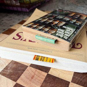 May include: Two cookbooks stacked on a wooden cutting board. The top book is titled "An Herb and Spice Cook Book" with a green bookmark that reads "Diamond Plates." The bottom book has a bookmark with the text "MAY" and "A CALIFORNIA LIVING BOOK."