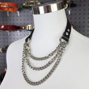 Puede incluir: Un collar gargantilla de cuero negro con tres hileras de cadenas plateadas. El collar tiene un anillo de metal plateado y se exhibe en un maniquí blanco. El fondo muestra otros collares de cuero.