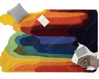 May include: A large, irregularly shaped area rug featuring a vibrant, multi-colored abstract pattern of flowing, layered bands in shades of yellow, orange, red, green, blue, and dark purple, with the word RUGABLES printed repeatedly across the surface.