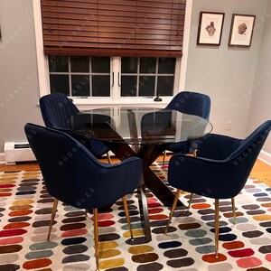 May include: A dining room set featuring a round glass table with a dark wood base and four navy blue velvet chairs with gold legs. The chairs surround a colorful patterned rug with an oval design. A window with brown blinds is visible.