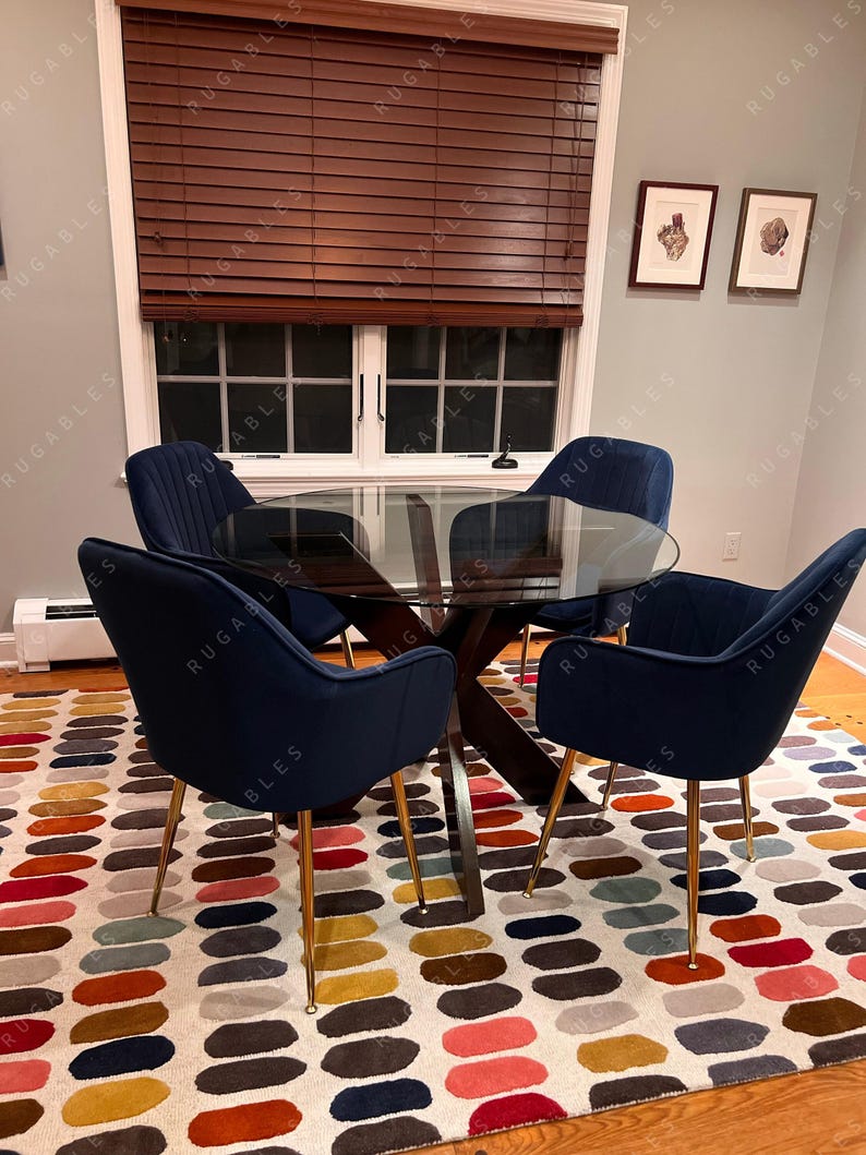 May include: A dining room set featuring a round glass table with a dark wood base and four navy blue velvet chairs with gold legs. The chairs surround a colorful patterned rug with an oval design. A window with brown blinds is visible.