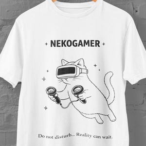 May include: White t-shirt featuring a black and white illustration of a cat wearing a VR headset and holding controllers. The text "+ NEKOGAMER +" and "Do not disturb... Reality can wait." are also printed on the shirt.