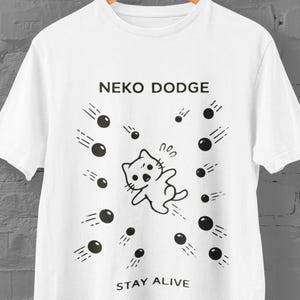 Könnte beinhalten: Weißes T-Shirt mit einer schwarzen Grafik einer Katze, die fallenden schwarzen Kugeln ausweicht. Der Text "NEKO DODGE" steht über der Grafik, und "STAY ALIVE" steht darunter. Das T-Shirt hängt an einem Holzkleiderbügel.