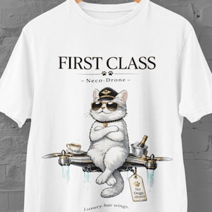 Pode incluir: Camiseta branca com um gato de desenho animado usando óculos de sol e chapéu de piloto, sentado em um drone. O texto "FIRST CLASS" e "Luxury has wings" são exibidos. Uma etiqueta diz "No Dogs Allowed".