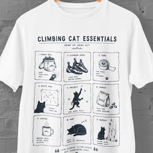 Peut inclure: T-shirt blanc avec un motif de chat grimpeur. Le design comprend des illustrations d'équipement d'escalade et un chat, avec le texte "Climbing Cat Essentials". Le t-shirt a un col rond et des manches courtes.