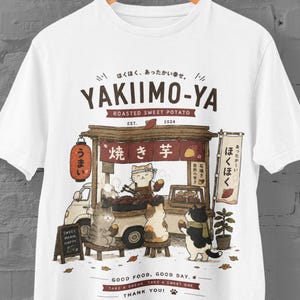 以下が含まれることがあります： 猫が運営する焼き芋屋台「YAKIIMO-YA」のグラフィックイラストがプリントされた白いTシャツ。日本語の文字、赤い提灯、小さな黒板、焼き芋を食べている3匹の猫が描かれています。