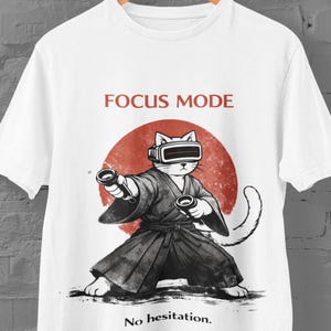 May include: White t-shirt featuring a cartoon cat in a samurai outfit wearing a VR headset and holding controllers. The design includes a red sun graphic and the text "FOCUS MODE" and "No hesitation."