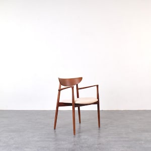 Può includere: Poltrona moderna di metà secolo con schienale e braccioli in legno curvi. La sedia ha un cuscino di seduta colorato chiaro ed è sostenuta da quattro gambe in legno sottili e angolate. Il legno ha un caldo tono marrone.