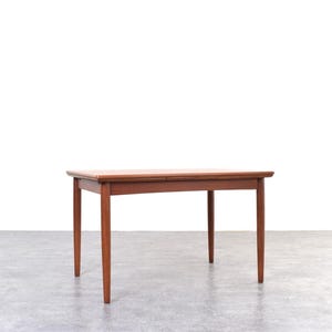 May include: A rectangular, mid-century modern wooden table with four tapered legs. The table is a warm reddish-brown colour and sits on a grey floor against a white background. The table's dimensions are approximately 122 cm long, 76 cm wide, and 74 cm high.