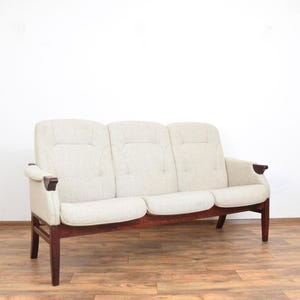 Op de afbeelding: Een driezitsbank met lichtbeige stof en een donkerhouten frame. De bank heeft een modern design uit het midden van de eeuw met een strakke, minimalistische esthetiek. De armleuningen hebben donkere houten accenten.