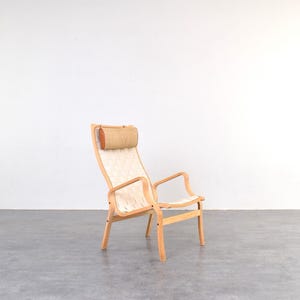 Può includere: Una poltrona da salotto con struttura in legno chiaro e seduta e schienale intrecciati. La sedia ha un poggiatesta e braccioli. La sedia è appoggiata a una parete bianca e un pavimento grigio.