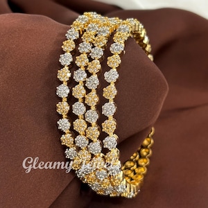 May include: Four gold and silver bangles with a floral design. The bangles are arranged on a brown surface. The text "Gleamy Jewels" is visible in the lower left corner.
