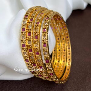 Peut inclure: Un ensemble de quatre bracelets rigides dorés présentant un motif métallique complexe avec des pierres carrées rouges et transparentes alternées. Les bracelets ont un design géométrique répétitif et le texte Gleamy Jewels est visible.