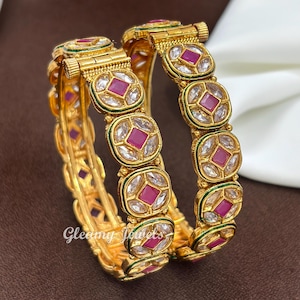 Peut inclure: Deux bracelets rigides dorés à charnière présentant un motif répétitif de pierres roses carrées entourées de pierres blanches transparentes dans un sertissage circulaire. Le métal inclut une charnière et le texte Gleamy Jewels.