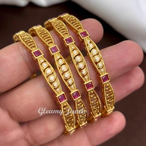 May include: Four gold-toned bangles featuring a textured metal design with white teardrop-shaped inlays and square-cut red stones. The bangles are arranged in a row and display the text Gleamy Jewels in white script across the lower section.