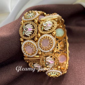 May include: Gold-toned bangles with intricate floral and pearl detailing. The bangles feature pink, white, and green accents, with the text "Gleamy Jewels" visible on the surface. The jewelry is set against a brown background.