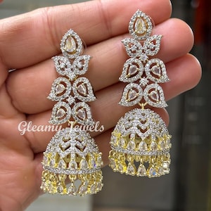 Könnte beinhalten: Ein Paar goldfarbene Ohrringe mit mehrschichtigem Design. Die Ohrringe haben klare, funkelnde Steine und einen glockenförmigen Boden mit hängenden Akzenten. Der Text "Gleamy Jewels" ist auf dem linken Ohrring sichtbar.