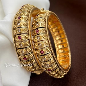 May include: Two gold-toned bangles featuring a textured, segmented design with alternating embedded clear and deep red stones. The surface of each bangle displays intricate floral-like metalwork patterns and the text Gleamy Jewels.