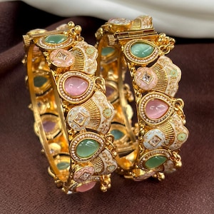 May include: Two gold bangles with detailed patterns. Each bangle has alternating pink and green teardrop-shaped gemstones, small square gems, and white enamel details. The bangles are displayed on a brown backdrop.