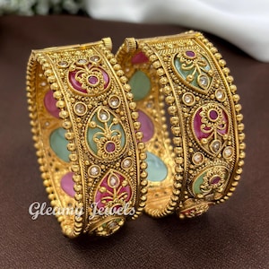 May include: Two gold-toned bangles featuring intricate metalwork, beaded edges, and alternating teardrop-shaped inlays in shades of pink and light green. Each inlay is centred with a small floral motif and clear stones. Text reads Gleamy Jewels.
