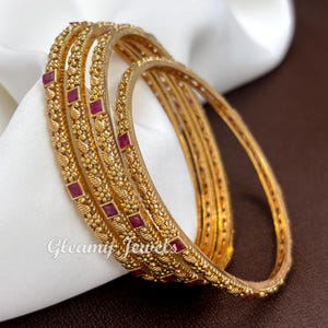 May include: A set of four gold-toned bangles featuring a textured, beaded surface and small, square-cut red stones embedded at regular intervals. The metal has a matte finish, and the text Gleamy Jewels is visible in white script on the lower left.