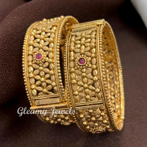 May include: Two gold-toned bangles featuring an intricate floral pattern with small, raised circular details and a single embedded red stone in the center of each. The metal surface has a textured finish, and the text Gleamy Jewels is visible.