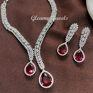 May include: A dazzling jewelry set featuring a necklace and matching earrings. The necklace and earrings are silver-toned and encrusted with sparkling clear stones. Each piece has a large, teardrop-shaped, deep red stone as a focal point. The text "Gleamy Jewels" is visible.