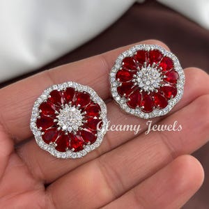 May include: A pair of floral-shaped stud earrings. Each earring features a central cluster of small, clear stones surrounded by teardrop-shaped red gemstones. The outer edge is lined with clear stones. The text "Gleamy Jewels" is visible.