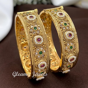 May include: A pair of gold-toned hinged bangles featuring intricate metalwork, embedded with small white pearls, clear stones, and alternating circular accents containing central red or green stones. The text Gleamy Jewels is visible at the bottom.
