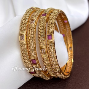May include: A set of three gold-toned bangles featuring a textured, granular surface and inlaid square-cut stones in shades of deep pink and clear white. The metalwork displays a repeating pattern of small beads, with the text Gleamy Jewels visible.