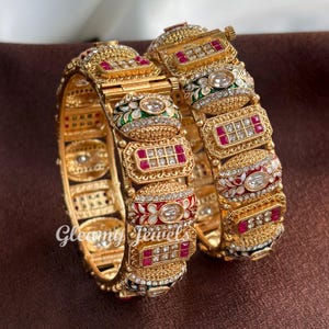 May include: Two ornate gold bangles with intricate detailing, featuring red, white, and green accents. The bangles are adorned with sparkling gemstones and a repeating rectangular pattern. The text "Gleamy Jewels" is visible on one of the bangles.