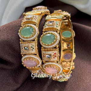 May include: Two gold bangles with oval green and pink gemstones, white enamel detailing, and small pearl accents. The bangles have a repeating pattern of gemstones and decorative elements. The text "Gleamy Jewels" is visible.