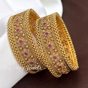 May include: Two gold-toned bangles featuring a textured, beaded border and an intricate central band. The design is embellished with small, clear, round stones and interspersed circular, deep pink stones. The text Gleamy Jewels is visible.