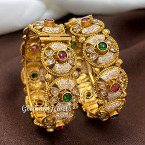 May include: Two gold-coloured bangles with intricate floral and geometric designs. Each bangle features white enamel, red, green, and clear gemstones. The bangles are ornate and detailed, with a vintage or antique aesthetic. The text "Gleamy Jewels" is visible.
