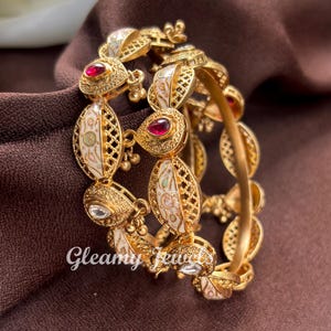 Könnte beinhalten: Goldfarbene Armreifen mit aufwendigen Filigranarbeiten und weißen Emaille-Details. Jeder Armreif hat ovale Akzente mit roten und klaren Edelsteinen. Der Text "Gleamy Jewels" ist unten links sichtbar.