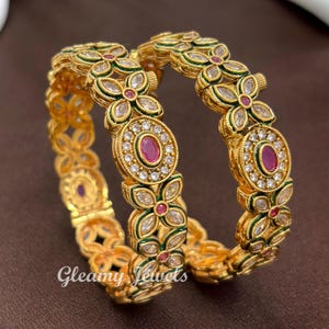 May include: Two gold-toned bangles featuring a repeating floral pattern with clear marquise-cut stones, small round white stones, and central oval-shaped pink stones. The metalwork includes green enamel accents and the text Gleamy Jewels.