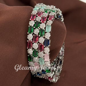 May include: Silver-toned bangles with a colorful array of gemstones. The bangles feature rectangular and round-cut stones in shades of red, green, blue, and pink, interspersed with smaller, clear stones. The text "Gleamy Jewels" is visible.