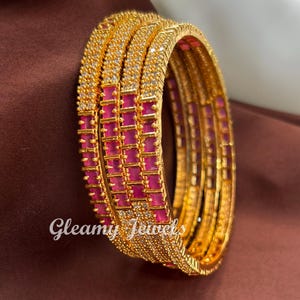 May include: A collection of gold-coloured bangles featuring a repeating pattern of pink rectangular stones. The top of the bangles are encrusted with small, clear stones. The text "Gleamy Jewels" is visible on the bottom left.