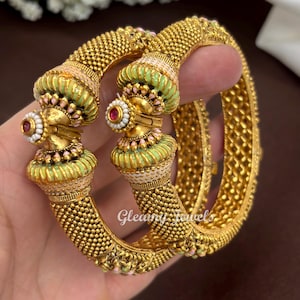 May include: Two gold-toned bangles featuring a textured beaded surface and ornate end caps decorated with light green and white enamel accents, small white beads, and a central red stone. The text Gleamy Jewels is visible on the bangles.