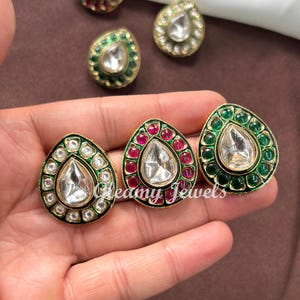 May include: Close-up of several ornate teardrop-shaped earrings. The earrings feature a central clear stone, surrounded by smaller stones in red, green, and white, set in gold-coloured metal. The earrings are displayed on a brown surface.