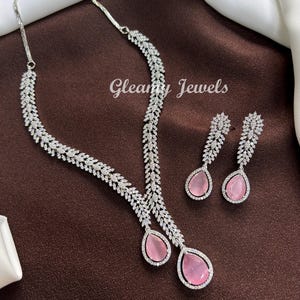May include: A silver necklace and earring set. The necklace and earrings are decorated with clear, sparkling stones and pink, teardrop-shaped gemstones. The brand name "Gleamy Jewels" is visible in the image.