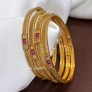 May include: A set of four gold-toned bangles featuring a textured beaded surface and alternating square-cut red and clear stones. The bangles have a metallic finish and the text Gleamy Jewels is visible on the lower left side of the image.