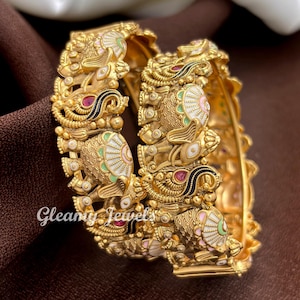 May include: Two gold-toned bangles featuring intricate embossed elephant motifs with white, pink, and green enamel detailing and small red accents. The metal surface has a textured finish, and the text Gleamy Jewels is visible on the left side.