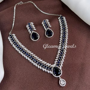 May include: A silver-toned necklace and earring set. The necklace and earrings feature dark blue teardrop-shaped stones and are accented with clear, sparkling stones. The necklace has a matching pendant. The text "Gleamy Jewels" is visible.