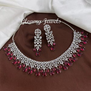May include: A silver necklace and earring set with clear and red gemstones. The necklace and earrings have a sparkling design with clear stones and teardrop-shaped, deep red gems. The jewellery is displayed on a brown surface.