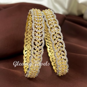 May include: Two gold-toned bangles with a leaf-like design, encrusted with clear gemstones. The bangles are set against a brown fabric background. The text "Gleamy Jewels" is visible on the left bangle.