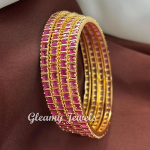 May include: A collection of gold bangles, each adorned with rows of bright pink, rectangular gemstones. The bangles are stacked, highlighting their detailed design and the sparkle of the gems. The image includes the text "Gleamy Jewels".