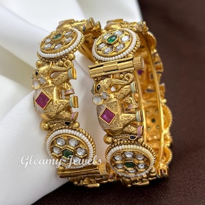 May include: A pair of gold-toned hinged bangles featuring embossed elephant motifs with pink square accents, surrounded by circular floral patterns inlaid with white stones and small green centre stones, finished with delicate white beaded borders.