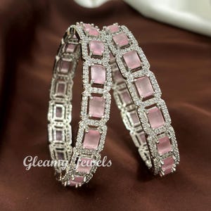 May include: Two silver bangles with rectangular pink gemstones and clear accent stones. The bangles are stacked together, with the text "Gleamy Jewels" in the lower left corner. The background is a solid brown colour.