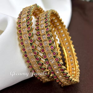 Peut inclure: Deux bracelets rigides dorés ornés d'une rangée centrale de pierres carrées transparentes, entourées de pierres roses circulaires et d'émail vert. Les bords métalliques présentent un motif torsadé. Le texte Gleamy Jewels est visible à gauche.